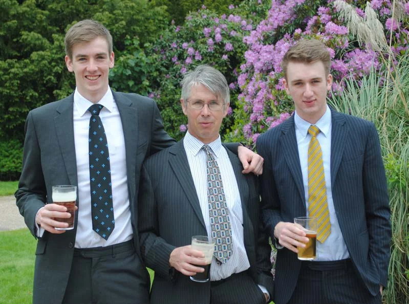 John Murphy in a suit standing between two taller young men, all holding drinks in a garden